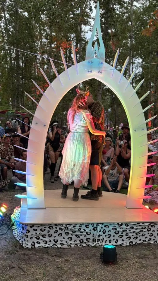 A couple kissing on the platform under the lit arch, guests watching from the grass